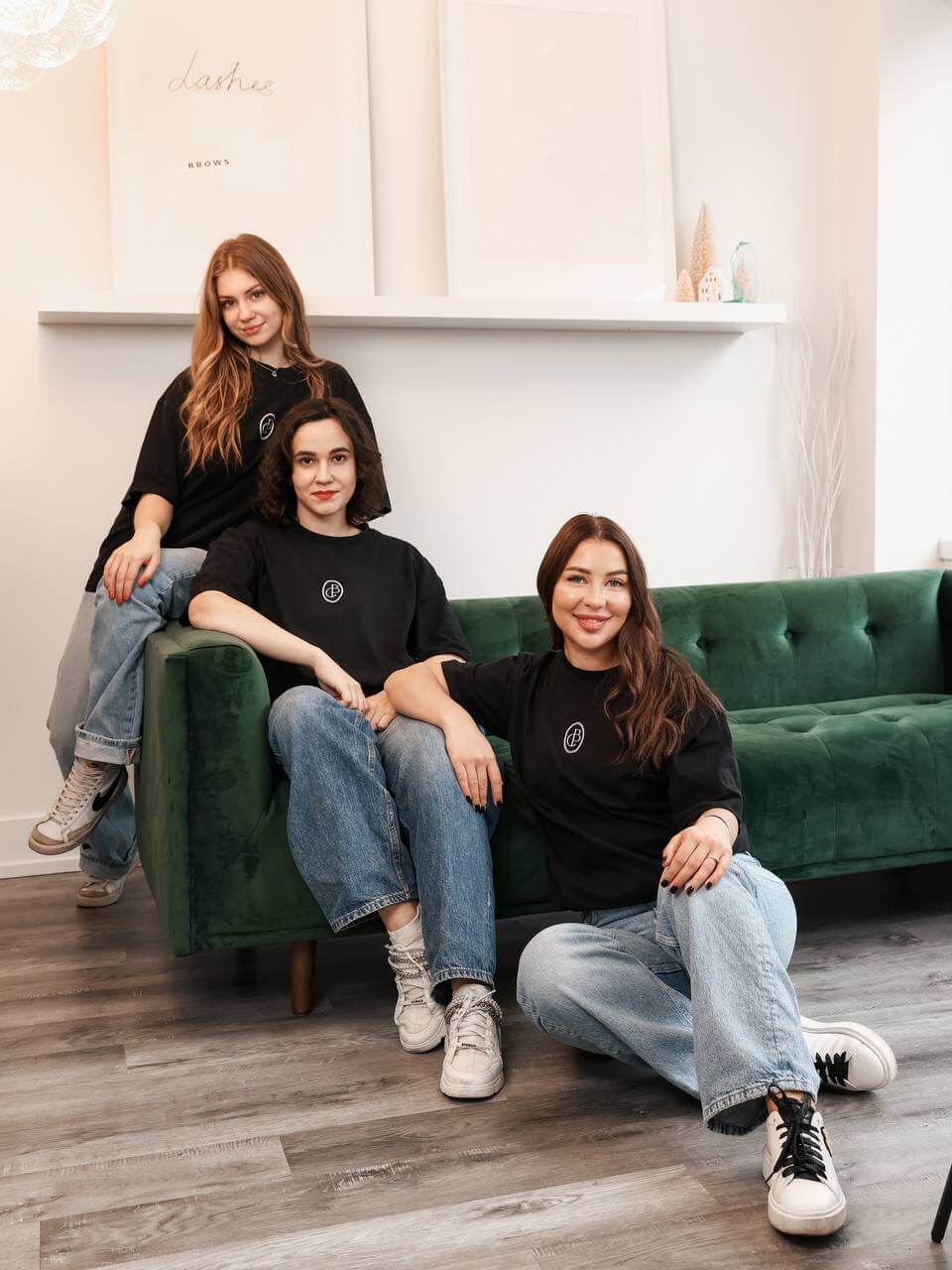 Three women in black shirts and jeans relax in a modern living room with wooden flooring, showcasing minimalist wall art. Two sit on the green sofa, while one leans against it, all effortlessly stylish as they discuss their latest eyelash extension experiences.