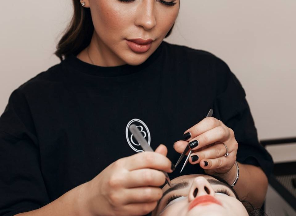 A woman with long dark hair is meticulously applying makeup to another womans eyebrows, while also delicately enhancing her look with eyelash extensions. The woman receiving the treatment is lying down with her eyes closed in a professional setting.